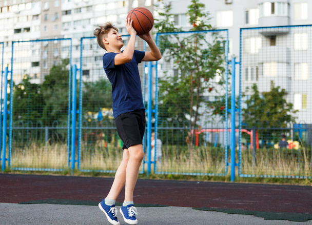Cute boy in blue t shirt plays basketball on city playground. Active teen enjoying outdoor game with orange ball. Hobby, active lifestyle, sport for kids. - Foto, imagen