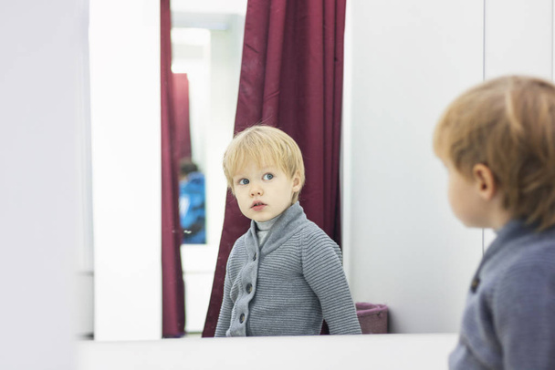 Serious Child - European blond boy tries on clothes in a store in the dressing room and looks at himself in the mirror. Concept of shopping, style and fashion. - Φωτογραφία, εικόνα