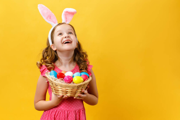 Beautiful cute little girl in Easter bunny ears holds a basket with eggs on a yellow background. The child raised his head and looks up. Copy space - Φωτογραφία, εικόνα