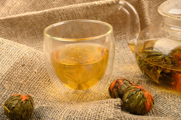 Blütentee gebrüht in einer gläsernen Teekanne, ein Glas Blütentee und Bällchen Blütentee auf einem Hintergrund aus grobem selbstgesponnenem Stoff. Nahaufnahme. - Foto, Bild