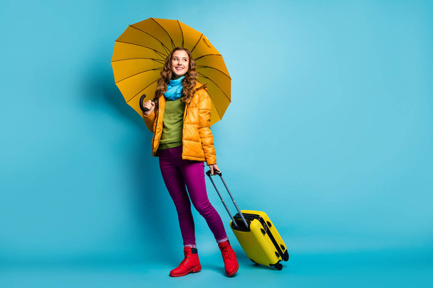 Full size photo of beautiful lady traveler abroad hold umbrella rolling bag walk airport flight wear yellow overcoat blue scarf trousers boots isolated blue color background - Foto, afbeelding