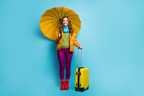 Full length photo of pretty traveler lady hold umbrella rolling bag suitcase wait airport journey wear yellow overcoat blue scarf violet trousers boots isolated blue color background - Foto, afbeelding
