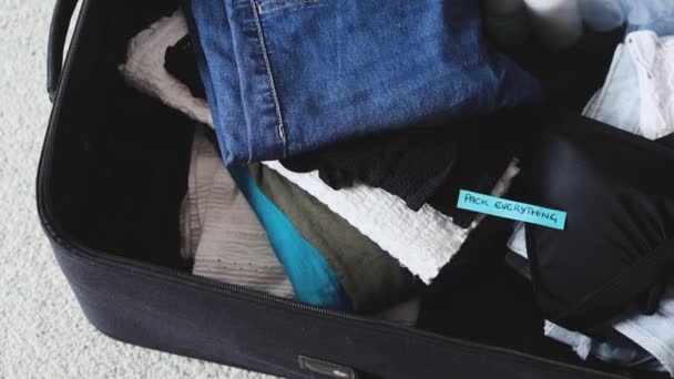 travel essentials for a holiday, luggage with pile of clothes and Packing Everything label on them with camera panning horizontally - Imágenes, Vídeo