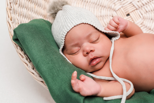 милый новорожденный смешанной расы ребенок в вязаной шляпе спит в корзине на белом
 - Фото, изображение