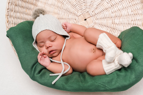  νεογέννητο μωρό μικτή φυλή σε πλεκτό καπέλο και κάλτσες ύπνου στο καλάθι σε λευκό - Φωτογραφία, εικόνα