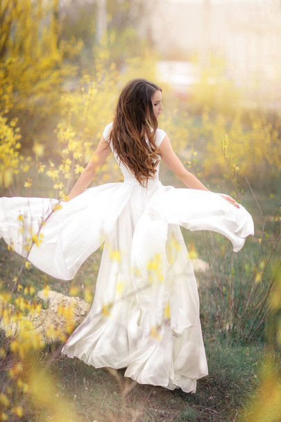Mujer en un vestido blanco en pleno crecimiento en un jardín con árboles con flores amarillas
 - Foto, Imagen