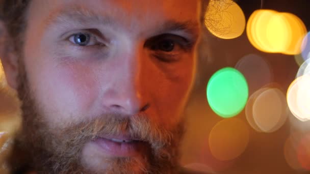 Close up, Portrait of a handsome Caucasian bearded long-haired young man looking straight and smiling on the background of moving evening lights of a city. - Footage, Video