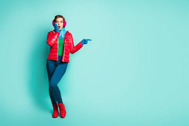 Full body photo of pretty lady indicate fingers empty space showing sale prices hand on cheek wear red overcoat blue scarf pink ear muffs pants shoes isolated teal color background - Photo, image
