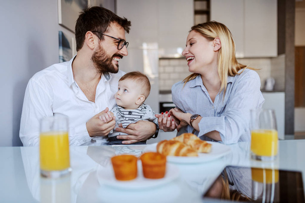 Young happy parents sitting in dining room with their adorable baby boy. Man holding baby. On table is breakfast. - Fotografie, Obrázek