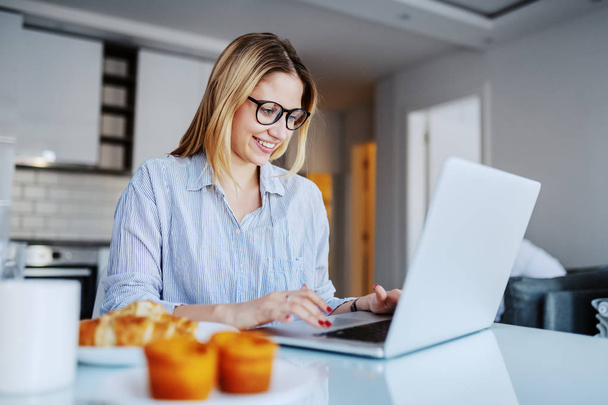 Cheerful caucasian blond woman with eyeglasses sitting at dining table in morning and using laptop. On table is breakfast. - Photo, Image