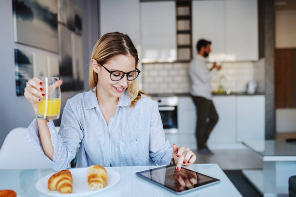 若い笑顔の白人女性がダイニングテーブルに座って眼鏡をかけ、新鮮なジュースのグラスを持ち、タブレットを使用しています。背景には夫が台所に立っていた。朝の時間. - 写真・画像