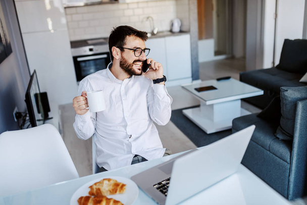 ハンサムな原因アジアのビジネスマンは、ダイニングテーブルに座ってカジュアルな服装で、新鮮な朝のコーヒーとマグカップを保持し、電話で話しています。テーブルの上にはノートパソコンと朝食. - 写真・画像