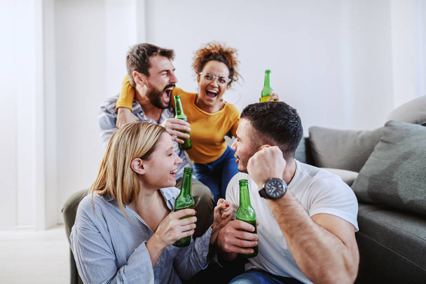 Grupo de amigos sentados en la sala de estar y animando a su equipo de fútbol favorito. Todos tienen botellas de cerveza.
. - Foto, imagen