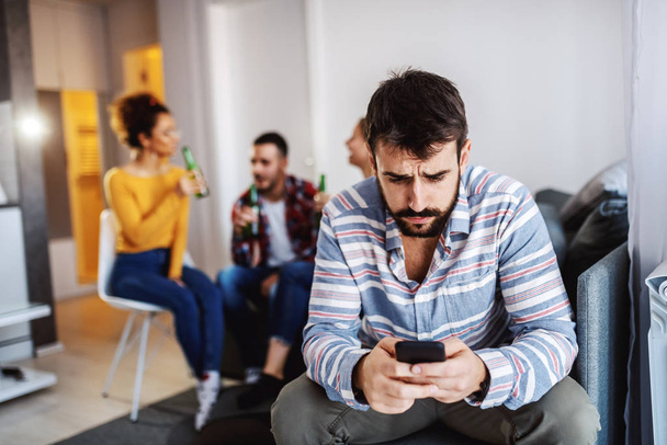 Attractive caucasian serious man in striped shirt using smart phone while his friends chatting and drinking in background. Living room interior. - Foto, imagen