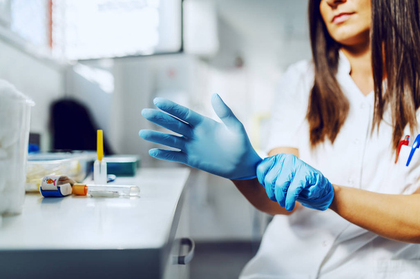 Обрезанная фотография кавказской привлекательной лаборантки в белой униформе, сидящей в лаборатории и надевающей стерильные резиновые перчатки
. - Фото, изображение