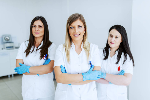 Trzech pięknych kaukaskich asystentów laboratoryjnych w białych mundurkach i gumowych rękawiczkach stojących w laboratorium z krzyżowanymi ramionami. - Zdjęcie, obraz