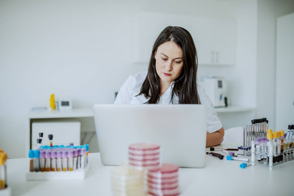 Beautiful caucasian female laboratory assistant sitting in lab and typing on laptop blood test results. On table are test tubes and petri dishes. - Foto, Imagen