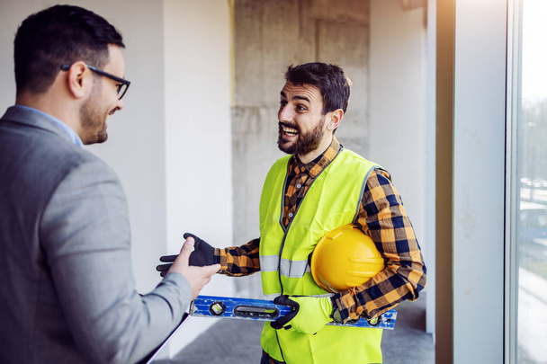 Handsome Caucasian bearded construction worker in work wear talking with his boss about project. Building in construction process interior. - Photo, Image