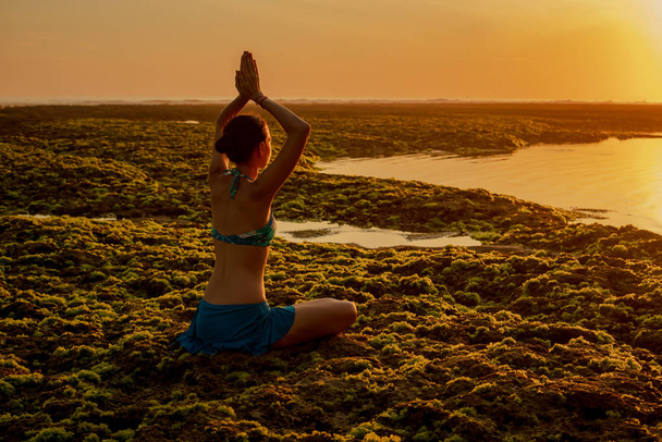 Mujer joven meditando, practicando yoga y pranayama en la playa. Práctica de yoga al atardecer. Levantando las manos en namaste mudra. Vista desde atrás. Playa de Melasti, Bali
 - Foto, Imagen