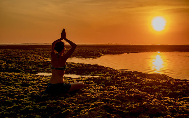 Jonge vrouw mediteren, yoga beoefenen en pranayama op het strand. Yoga bij zonsondergang. Handen omhoog in namaste mudra. Zicht van achteren. Strand van Melasti, Bali - Foto, afbeelding