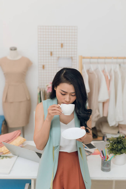 Happy young Asian fashion designer business drinking coffee at her workshop/store - Foto, imagen