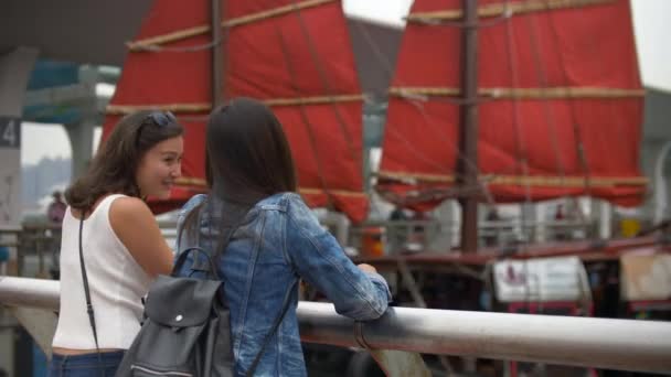 junge Frauen unterhalten sich lachend am Pier und beobachten das Boot mit den roten Segeln  - Filmmaterial, Video