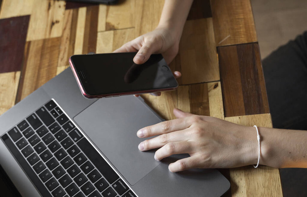 Τα χέρια των γυναικών χρησιμοποιούν ένα smartphone με φορητό υπολογιστή. Μελέτη και εργασία σε απευθείας σύνδεση, ελεύθερος επαγγελματίας. Αυτοαπασχολούμενη ή ανεξάρτητη γυναίκα, κορίτσι που εργάζεται με το laptop της κάθεται σε ξύλινο τραπέζι με τηλέφωνο και ηλεκτρονικό αναγνώστη. - Φωτογραφία, εικόνα