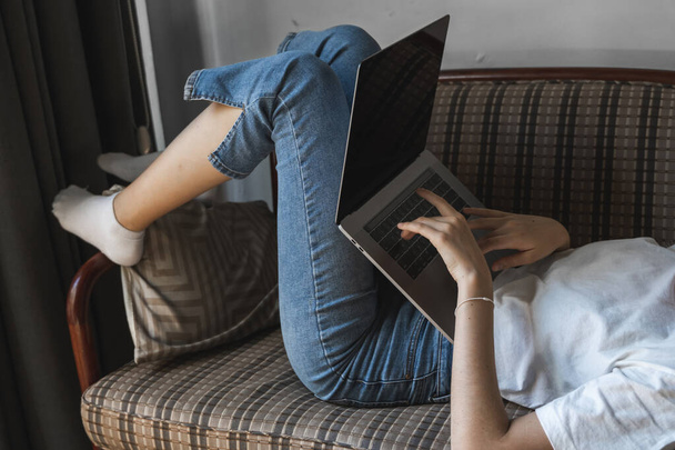 Γυναίκα με φορητό υπολογιστή ξαπλωμένη σε καναπέ. Μελέτη και εργασία σε απευθείας σύνδεση, ελεύθερος επαγγελματίας. Αυτοαπασχολούμενη γυναίκα, κορίτσι που εργάζεται με το σημειωματάριό της ξαπλωμένη σε έναν καναπέ με τηλέφωνο, smartphone και ηλεκτρονικό αναγνώστη στο τραπέζι. - Φωτογραφία, εικόνα