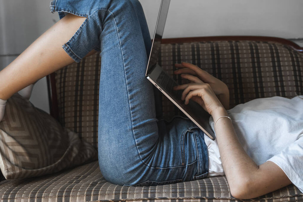 Woman with a laptop laying on a sofa. Study and work online, freelance. Self employed woman, girl working with her notebook laying on a couch with a phone, smartphone and ereader on table. - Foto, Bild