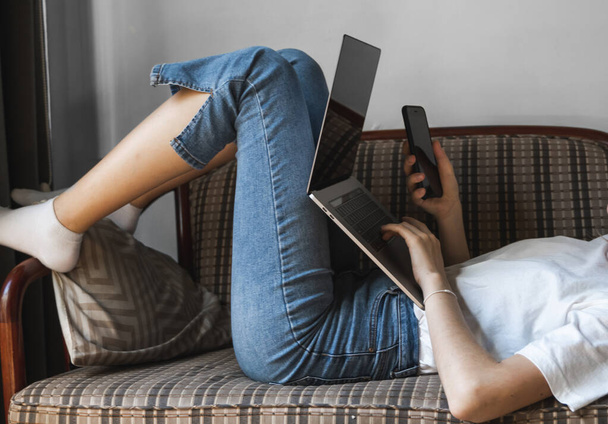 Woman with a laptop is laying on a sofa and using a smartphone. Study and work online, freelance. Self employed girl working with her notebook laying on a couch with a phone. - 写真・画像