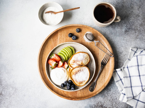 Delicious breakfast - fritters pancakes, sprinkled with powdered sugar with fruits and berries and sour cream on plate and cup of coffee on wood tray. Top view - Photo, Image