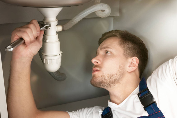 man plumber in uniform fixing the sink pipe with adjustable spanner in his hand lying on the kitchen floor looks concentrated professional plumbing repair service - Foto, immagini