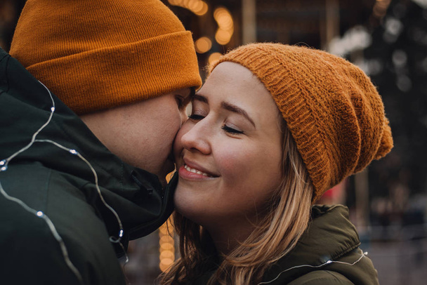 liebende schöne Paar in orangefarbenen Hüten und grünen Jacken umarmen und küssen sich auf dem verschwommenen Hintergrund mit Lichtern. Winterurlaub romantische Stimmung. Valentinstag-Konzept. - Foto, Bild