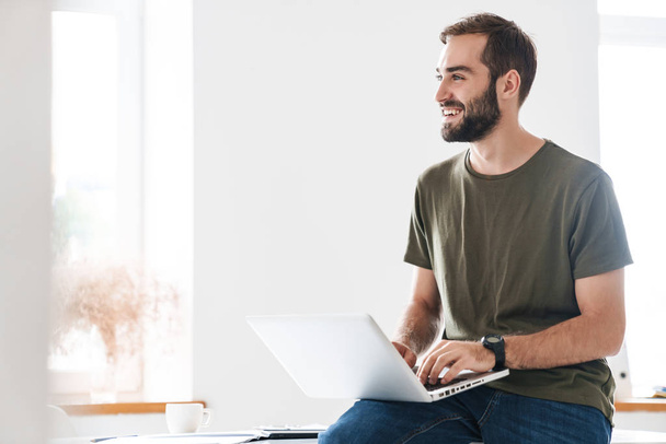 Image of handsome pleased man typing on laptop and laughing - Photo, image