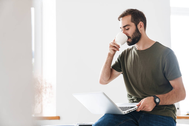 Image of handsome pleased man typing on laptop and drinking coffee - Fotó, kép