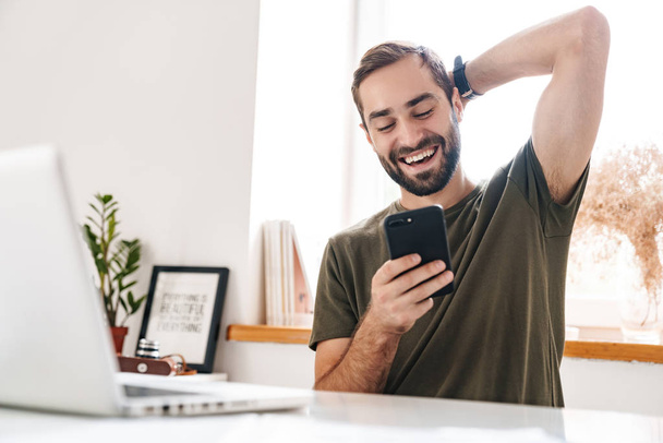 Obraz przystojnego śmiejącego się mężczyzny piszącego na laptopie i korzystającego z telefonu komórkowego - Zdjęcie, obraz