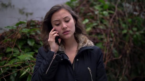 Hyvin järkyttynyt nuori sekoitettu nainen puhuu puhelimessa ulkona talvitakki päällä
 - Materiaali, video
