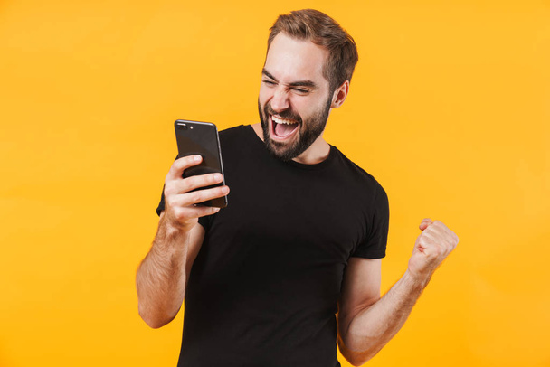 Image of happy man wearing t-shirt rejoicing and holding smartph - Foto, afbeelding