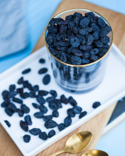 Delicious black raisins in a transparent glass on a white tray and a blue background. - Foto, imagen