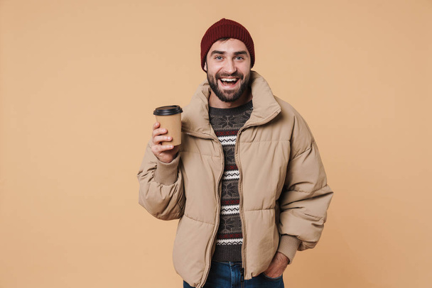 Image of young man in winter jacket and hat smiling while drinki - Photo, image