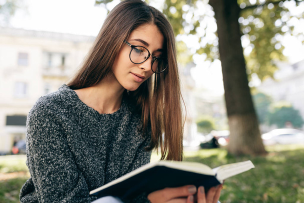 Smart young woman wearing sweater and transparent eyeglasses, sitting outdoor on the city street and reading the book while waiting her friends. Young female student learning in the city street.  - Photo, Image