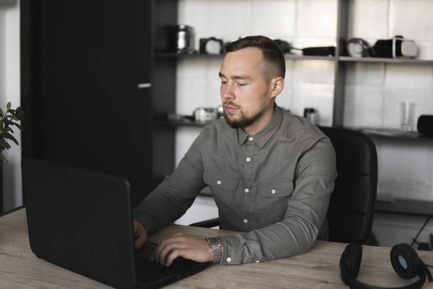 Young businessman or student in a shirt sitting against monitor of computer. Working on a pc at a table in the office with a thoughtful expression. Young businessman working on his laptop in office. - 写真・画像