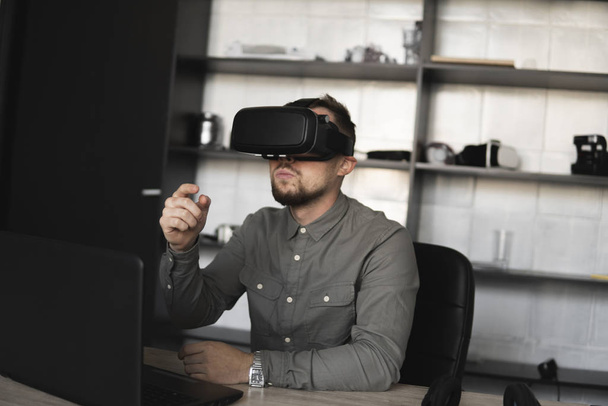 Young bearded man sitting against computer with virtual reality glasses trying to touch something or tap some thing in front of what he seeing. Modern technologies. The concept of future technology. - Fotografie, Obrázek