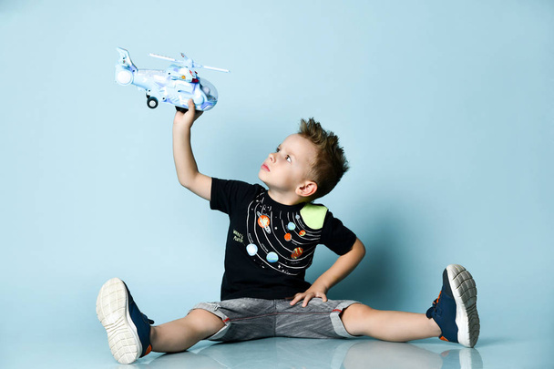 lächelnder blonder Junge in stylischer Freizeitkleidung, der mit ausgebreiteten Beinen auf dem Boden sitzt und einen Spielzeughubschrauber in der Hand hält - Foto, Bild