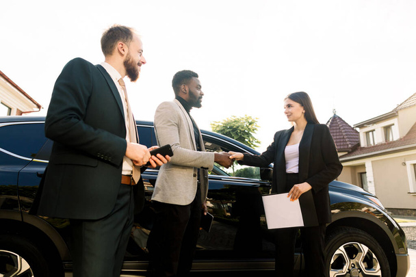 Handshake of a client, African young businessman and salesperson, young Caucasian woman at the yard of car showroom outdoors. Two men business partners buying new car - Фото, изображение