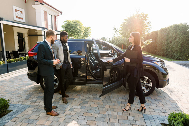 Pretty car saleswoman in business wear helping to young two multiethnical businessmen clients to make decision showing a new car, opening car doors, while standing on the yard of car salon outdoors - Photo, Image