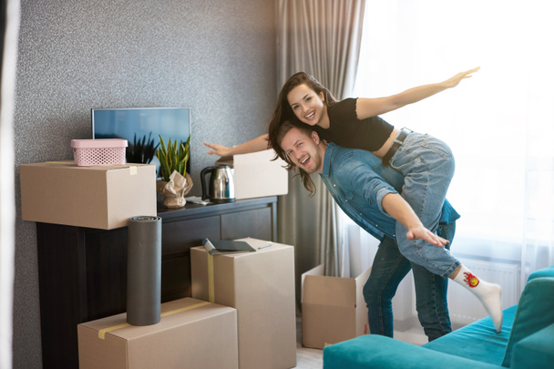 young couple man and woman looking happy while unpacking boxes , wife sitting on husband's shoulders, moving process - Photo, Image