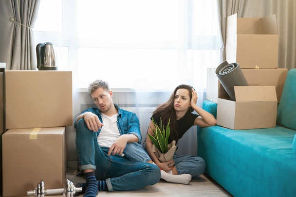 νεαρός άνδρας ζευγάρι και η γυναίκα αναζητούν κουρασμένος κάθεται στο πάτωμα κατά τη διάρκεια της μετάβασης σε νέο διαμέρισμα αποσυσκευασία κουτιά,, διαδικασία κίνησης - Φωτογραφία, εικόνα