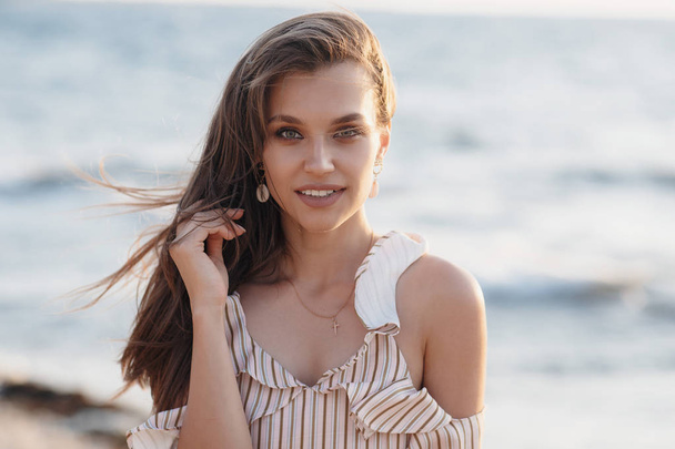 Beautiful young woman posing against the sea and skyline.Summer vacation concept. Happy woman relaxing near the sea on beach.Hipster slim girl in beige dress standing and tanning on beach near sea with waves,sunny warm weather. Peaceful calm moment. - Valokuva, kuva