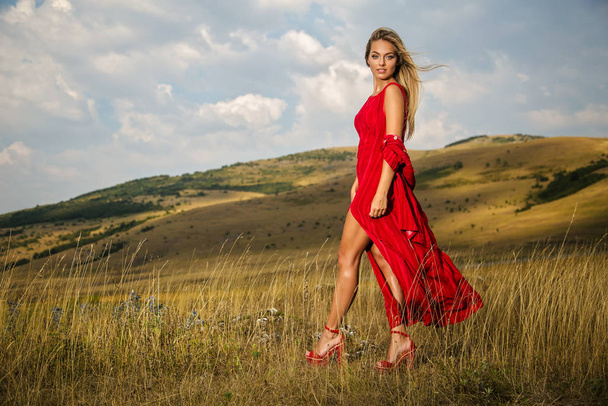 Une belle blonde en robe rouge estivale traverse un champ de montagne
 - Photo, image